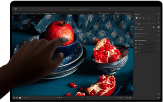 MacBook Pro mit PROLED XDR Display mit einem Stillleben mit Granatäpfeln, blauer Keramik und Textilien, eine Hand reicht nach dem Bildschirm, um mit dem Bild zu interagieren